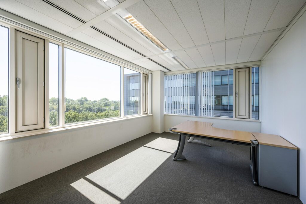 Lichte, lege kantoorruimte aan de Thomas R. Malthusstraat met groot hoekbureau en uitzicht op bomen en aangrenzend kantoorgebouw.